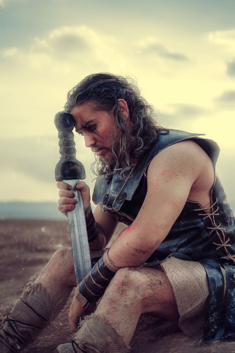 A pensive warrior in ancient armor holds a sword, seated outdoors during dusk.